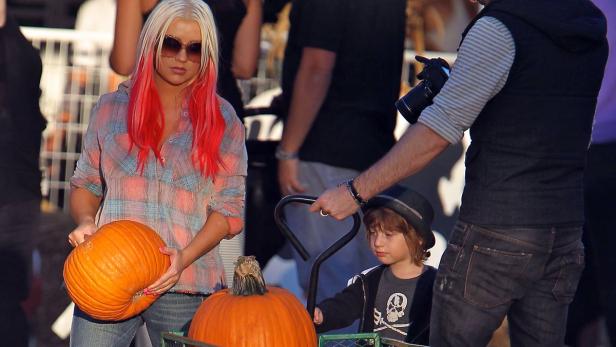 Christina Aguilera mit rotem Haar hält einen Kürbis, während ein Kind in einem Wagen sitzt.
