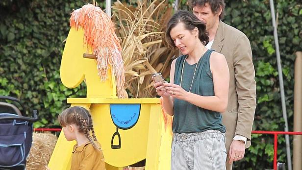Milla Jovovich mit Familie auf einem Kürbisfeld, sie schaut auf ihr Handy.