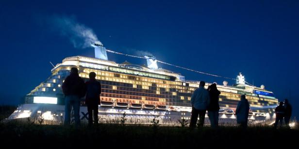 Eine Gruppe von Menschen betrachtet nachts ein großes, hell erleuchtetes Kreuzfahrtschiff.