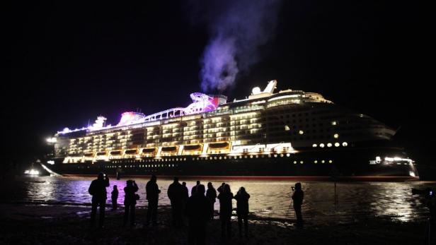 Ein großes Kreuzfahrtschiff liegt nachts vor Anker, während Menschen am Ufer zusehen.