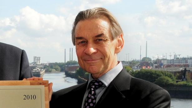 Ein Mann im Anzug hält eine Plakette mit der Aufschrift „2010“ vor einer Stadtlandschaft.