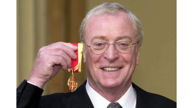 Michael Caine hält eine Medaille mit rotem Band in der Hand und lächelt.