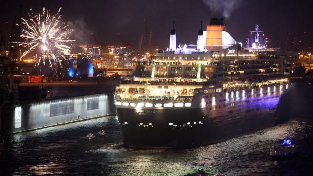 Die „Queen Mary 2“ fährt nachts in einen Hafen, während Feuerwerk den Himmel erhellt.