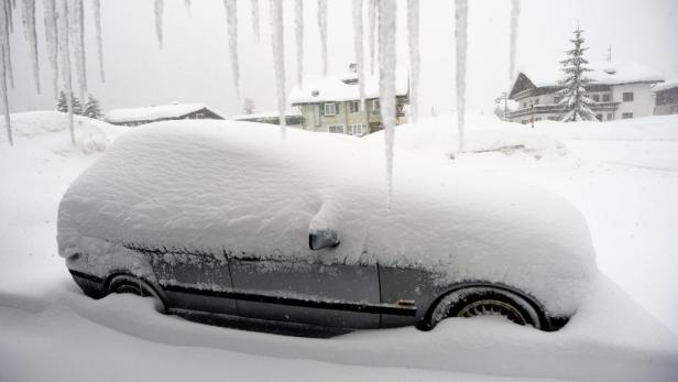 Ein Auto ist vollständig mit Schnee bedeckt, Eiszapfen hängen von einem Dach herab.