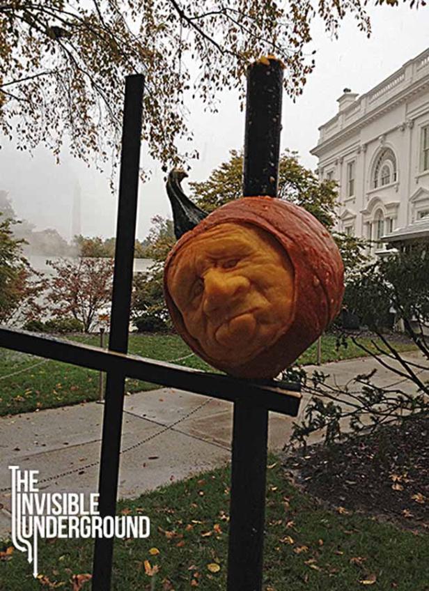 Ein geschnitzter Kürbis mit einem Gesicht steht auf einem Zaun vor einem Gebäude.