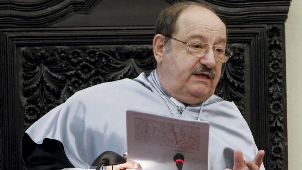 Ein Mann in akademischer Kleidung hält ein Dokument vor einem verzierten Hintergrund.