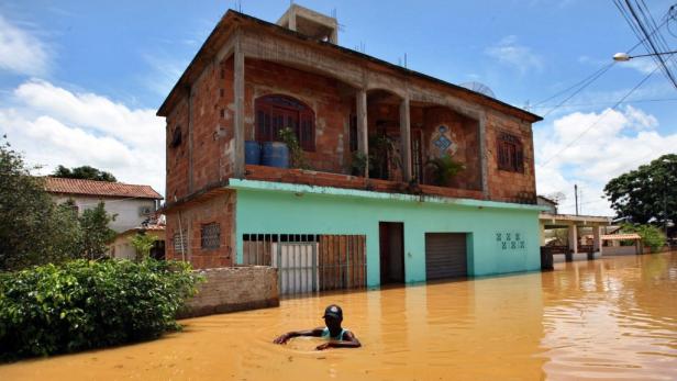 Ein Mann watet durch das braune, überflutete Wasser vor einem Haus.