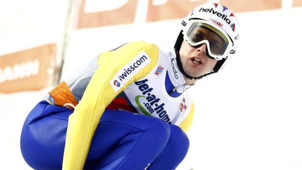 Ein Skispringer in gelb-blauer Kleidung und Helm mit Sponsorenlogos in der Hocke.