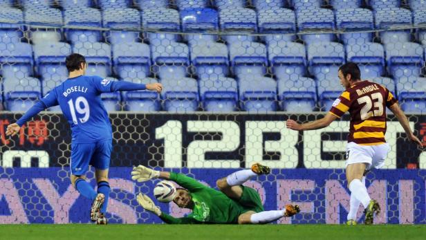 Ein Fußballtorwart wehrt einen Schuss ab, während zwei Spieler zusehen.