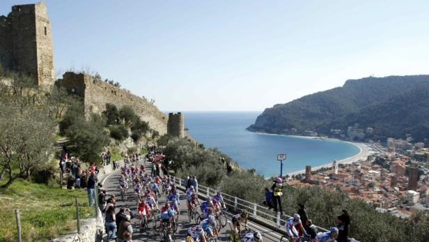 Ein Radrennen führt an einer alten Burgruine vorbei, mit Blick auf eine Küstenstadt.