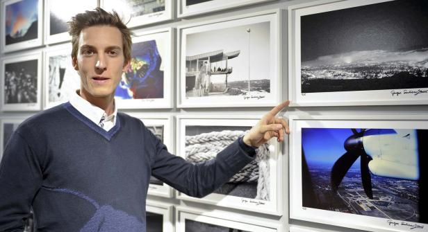 Ein Mann steht vor einer Wand mit gerahmten Fotografien und zeigt auf eines der Bilder.