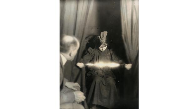 Eine junge Frau sitzt vor einem Vorhang und scheint einen Lichtblitz zu erzeugen, während ein Mann zusieht.