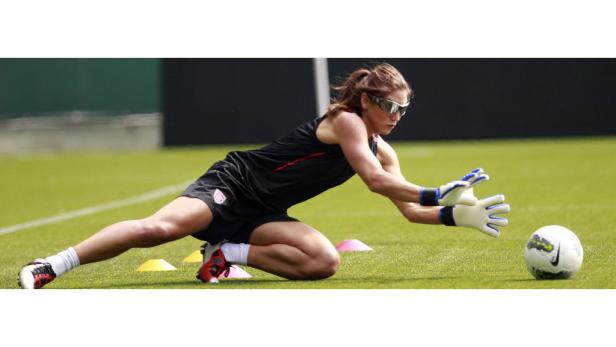 Eine Torhüterin mit Sonnenbrille hechtet nach einem Fußball.