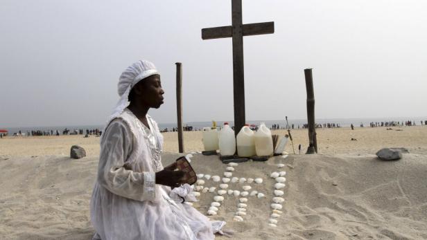 Eine Frau in weißer Kleidung betet vor einem Kreuz am Strand.