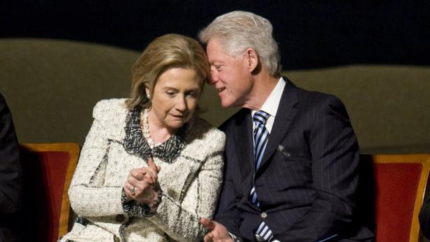 Bill und Hillary Clinton sitzen nebeneinander, während er ihr etwas ins Ohr flüstert.
