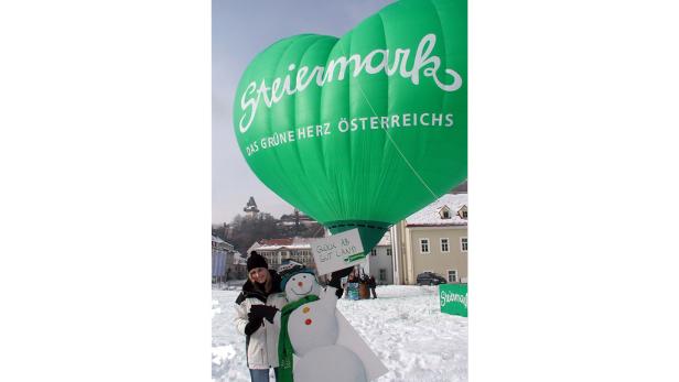 Eine Frau posiert mit einem Schneemann vor einem grünen Heißluftballon mit der Aufschrift „Steiermark“.