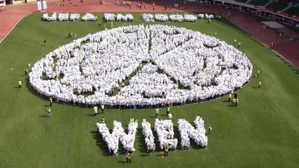 Viele Menschen in weißen T-Shirts bilden ein Peace-Zeichen und das Wort „Wien“ auf einem Rasen.