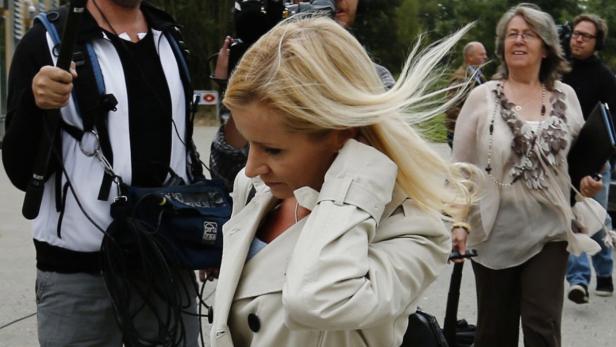 Eine blonde Frau in einem Trenchcoat geht an einer Gruppe von Menschen und Kameraleuten vorbei.