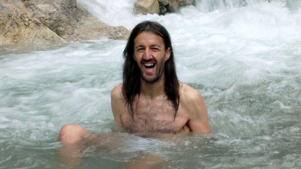 Ein Mann mit langen Haaren sitzt lachend in einem Fluss.