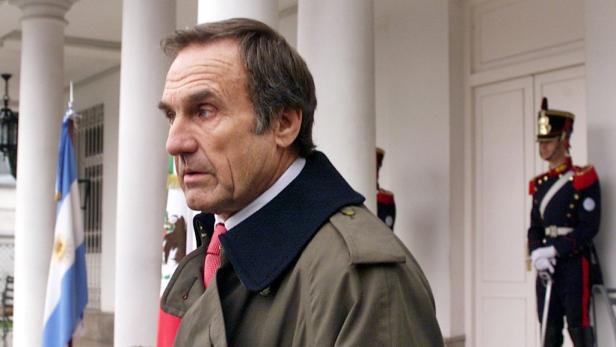 Ein Mann in einem Mantel steht vor einem Gebäude mit argentinischen Flaggen und einem Wachmann.