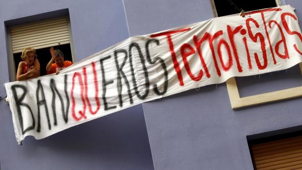 Zwei Frauen halten ein Transparent mit der Aufschrift „BANQUEROS Terroristas“ aus einem Fenster.