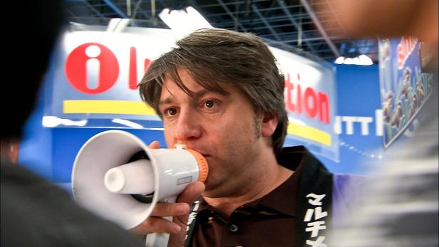 Ein Mann spricht durch ein Megaphon vor einem Stand mit der Aufschrift „Innovation“.