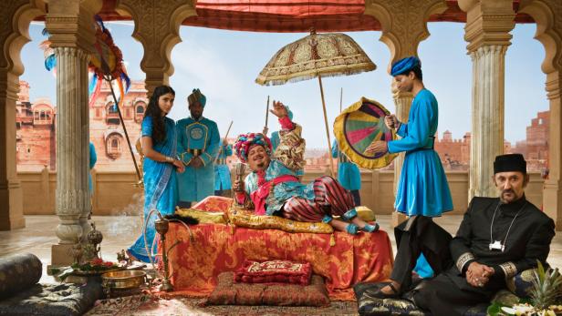 Ein Mann in prächtiger Kleidung liegt auf einem Sofa, umgeben von Dienern und einer Frau.