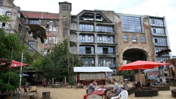 Ein Paar sitzt an einem Tisch im Freien vor einem Gebäude mit Graffiti in Berlin.