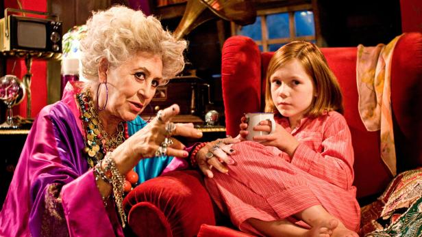 Eine ältere Frau mit Schmuck gestikuliert, während ein junges Mädchen mit einer Tasse auf einem roten Sessel sitzt.