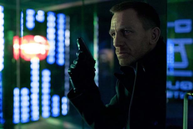 Daniel Craig als James Bond mit einer Waffe in der Hand vor Neonlichtern.