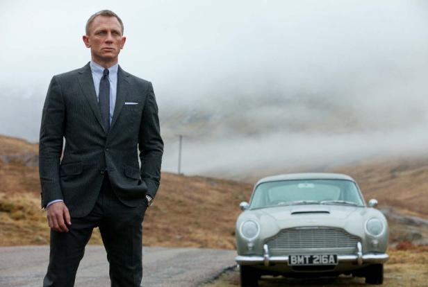 Daniel Craig als James Bond posiert neben einem Aston Martin in einer nebligen Landschaft.