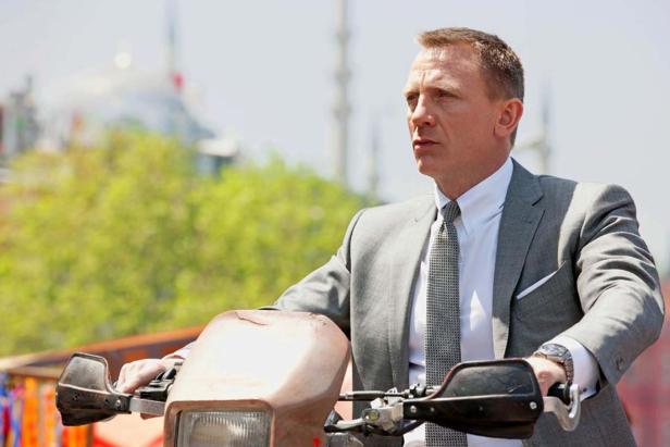 Daniel Craig fährt in einem grauen Anzug einen Motorroller.