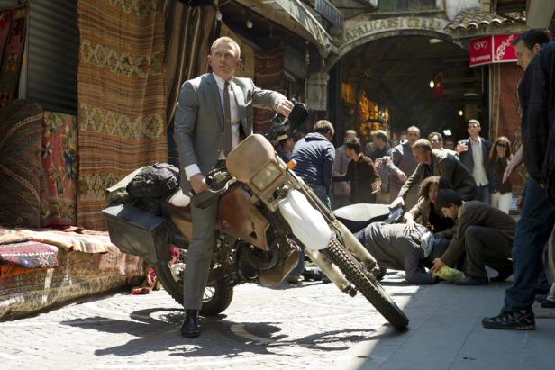 Daniel Craig sitzt in einem grauen Anzug auf einem Motorrad in einer belebten Gasse.