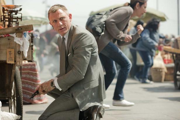 Daniel Craig als James Bond kniet mit einer Waffe auf einem belebten Marktplatz.