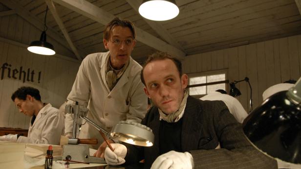 Zwei Männer in Arbeitskleidung sitzen an einem Tisch mit Dokumenten und Werkzeugen.