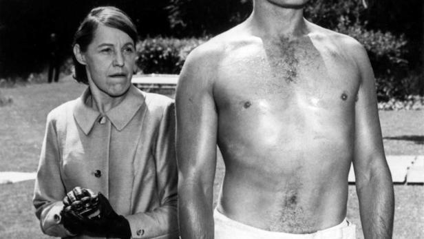 Eine Frau in Mantel blickt auf einen verschwitzten, barbusigen Mann.