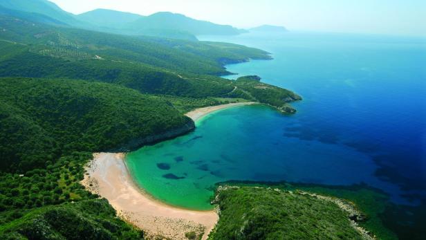 Blick auf eine malerische Bucht mit türkisfarbenem Wasser und einem Sandstrand, umgeben von grünen Hügeln.