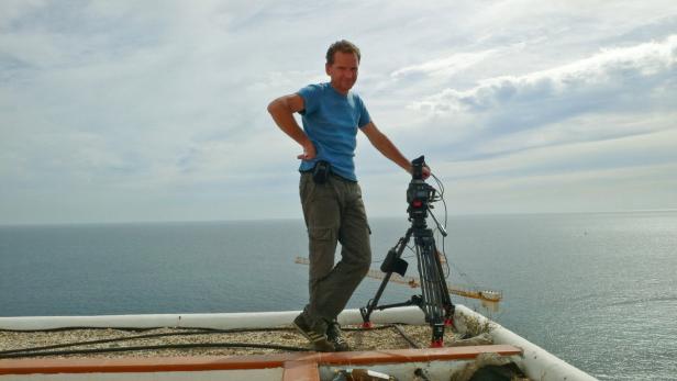 Ein Mann steht mit einer Kamera auf einem Dach mit Blick auf das Meer.