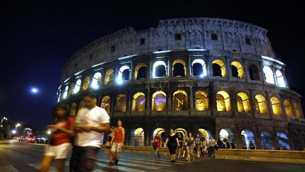 Das Kolosseum in Rom ist nachts beleuchtet, während Menschen die Straße überqueren.