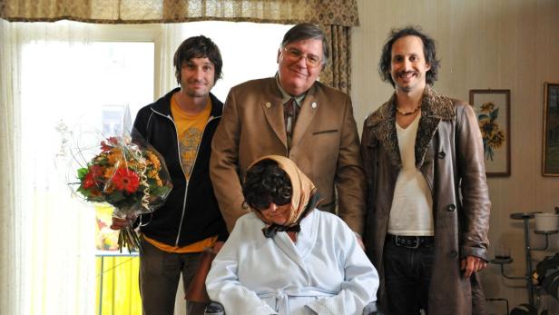 Eine Frau im Rollstuhl mit drei Männern im Hintergrund, einer davon mit einem Blumenstrauß.