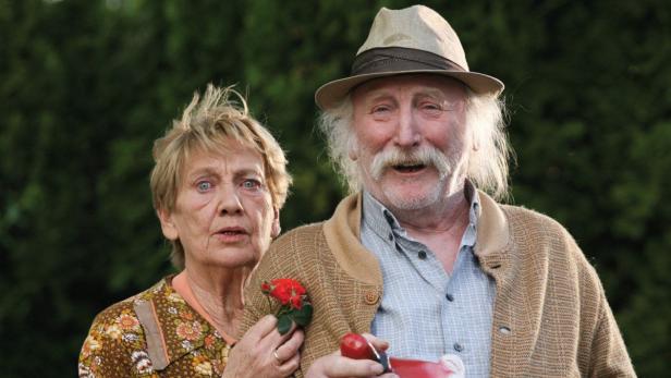 Ein älteres Paar steht vor einem grünen Hintergrund, der Mann hält eine rote Rose.