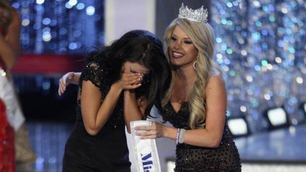 Eine gekrönte Frau tröstet eine weinende Frau mit einer Schärpe mit der Aufschrift „Miss“.