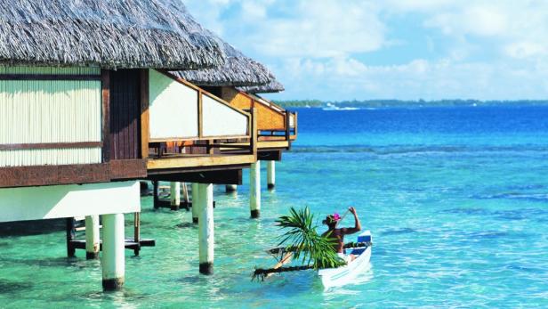 Eine Frau paddelt in einem Auslegerkanu an einem strohgedeckten Bungalow vorbei.