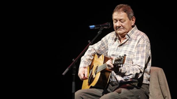 Ein älterer Mann sitzt auf einem Stuhl und spielt Gitarre.