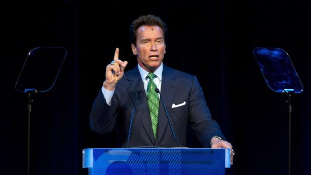 Arnold Schwarzenegger spricht auf einer Bühne und hebt warnend den Finger.