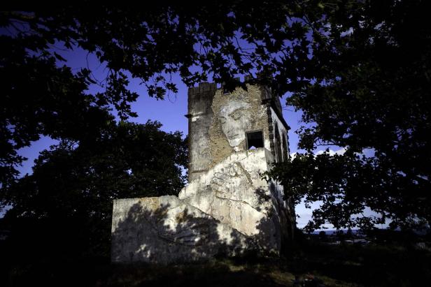 Eine alte, teilweise zerstörte Turmruine, umrahmt von Bäumen, vor blauem Himmel.