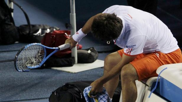 Ein Tennisspieler sitzt erschöpft auf einer Bank und hält ein Handtuch und einen Schläger.