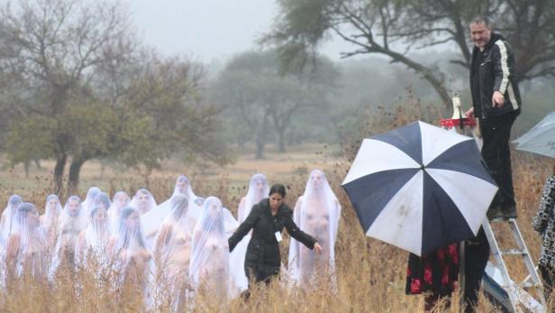 Eine Gruppe von Menschen, gehüllt in weiße Tücher, steht auf einem Feld; eine Frau und ein Mann mit Regenschirm sind ebenfalls zu sehen.