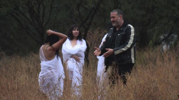 Ein Mann in Jacke gestikuliert zu drei Frauen in weißen Tüchern auf einer Wiese.