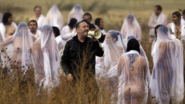 Ein Mann mit Megafon steht vor einer Gruppe von Menschen, die mit durchsichtigen Tüchern bedeckt sind.